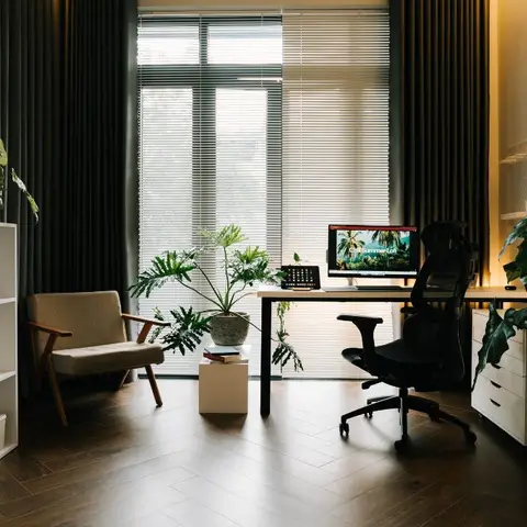 A compact home study with a black-frame desk against tall curtained windows, a mid-century armchair, white cube shelving, and layered potted plants.