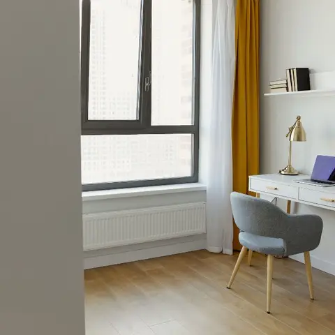 A compact study nook with a white writing desk, grey bouclé chair and mustard curtain against a floor-to-ceiling window overlooking a high-rise skyline.