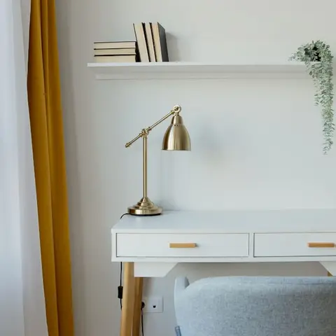 A compact white writing desk with brass tapered legs under a wall shelf of books, a brass desk lamp and a trailing plant beside a mustard curtain.