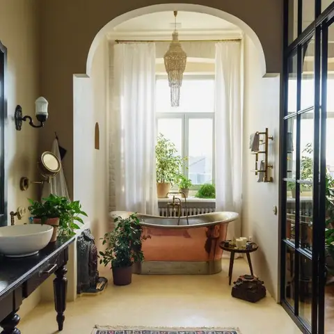 A copper freestanding tub sits under an arched doorway with a beaded chandelier and sheer curtains, opening into a glass-partitioned lounge area.