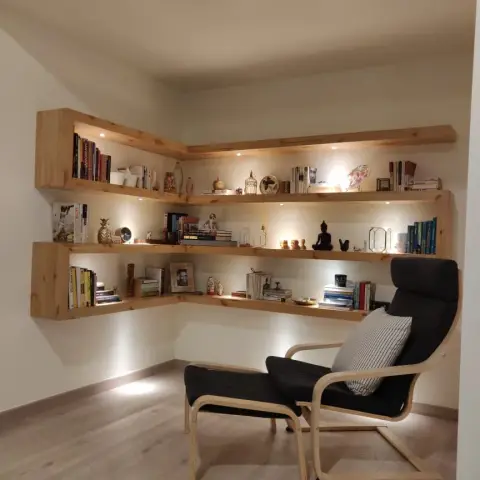 A corner reading nook with L-shaped floating wood shelves lit from beneath, styled with books and curios, paired with a black bentwood lounge chair and ottoman