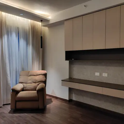 A cove-lit corner with a tan recliner beside floor-to-ceiling curtains, facing a wall-mounted TV console with a textured stone-look backsplash.