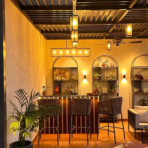 A covered terrace bar with arched metal display shelving, brass-trimmed pergola ceiling, and woven bar stools lit by warm pendant lanterns.