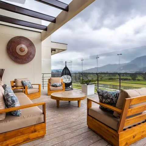 A covered terrace with chunky wood-frame outdoor sofas and armchairs around a round stone-top coffee table, glass railing and a mountain view