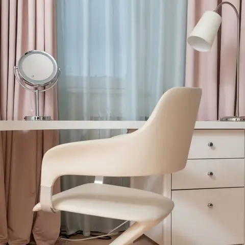 A cream desk nook with a white curved-back chair, a round vanity mirror and a task lamp framed by dual-layer blush and soft-blue curtains.