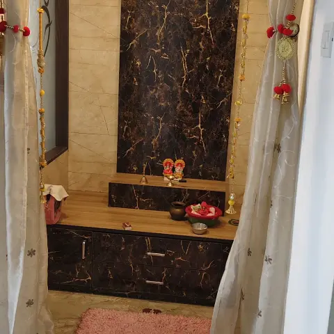 A curtained pooja alcove with a dark gold-veined marble backpanel, a wood-topped storage unit with deities and puja items