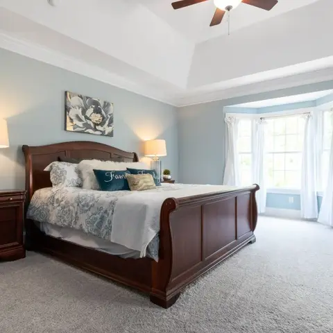 A dark cherry wood sleigh bed with matching nightstands under a tray ceiling, flanked by a bay window with sheer and drapery curtains.