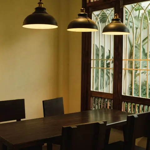 A dark wood dining table and chairs under three matching black dome pendant lights, tall arched wood-framed windows overlooking a lush garden