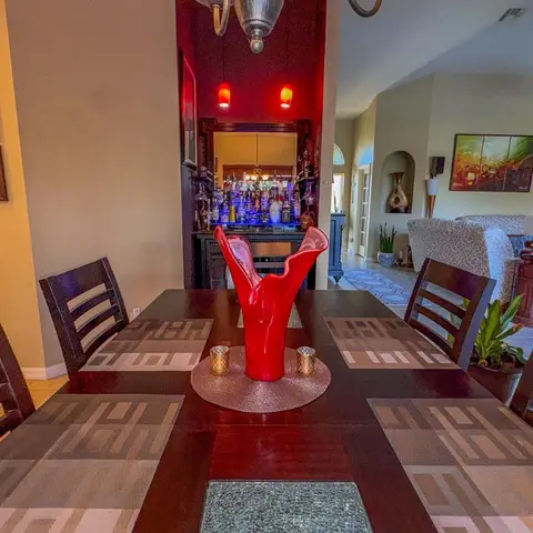 A dark wood dining table set with patterned placemats and a red sculptural vase, facing a mirrored home bar nook lit by red pendant lights.