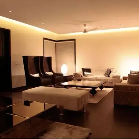 A dimly lit living room with dark wood flooring, tall wingback accent chairs, and a beige sectional sofa around a glossy black coffee table