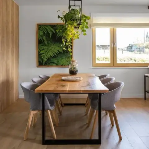 A dining nook with a live-edge wood table on black steel legs, grey upholstered chairs, a framed fern print, and a trailing pothos hanging above.