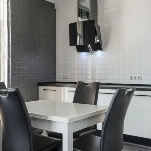 A dining nook with a white table and black leather chairs in front of a white subway-tile kitchen backsplash and an angular black chimney hood