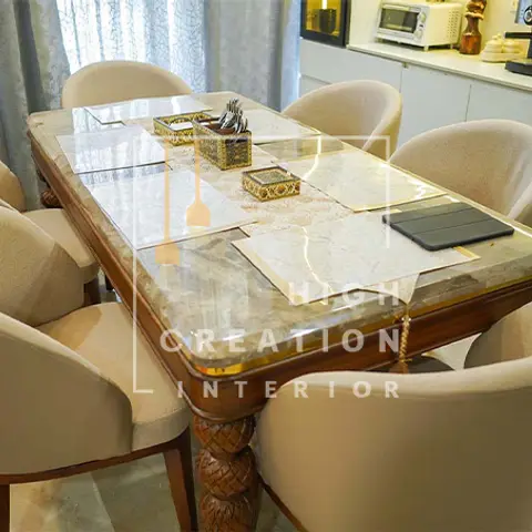A dining room with a carved wood dining table topped in marble, surrounded by six beige upholstered chairs, beside a white kitchen counter.