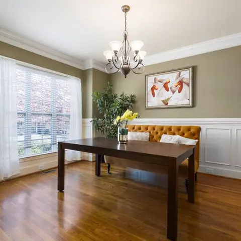 A dining room with a dark wood table and a mustard tufted bench beneath a five-arm chandelier, olive green walls with white wainscoting