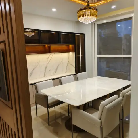 A dining room with a marble-top table under a crystal chandelier set into a gold mirrored ceiling feature, beside a black-and-marble crockery unit.