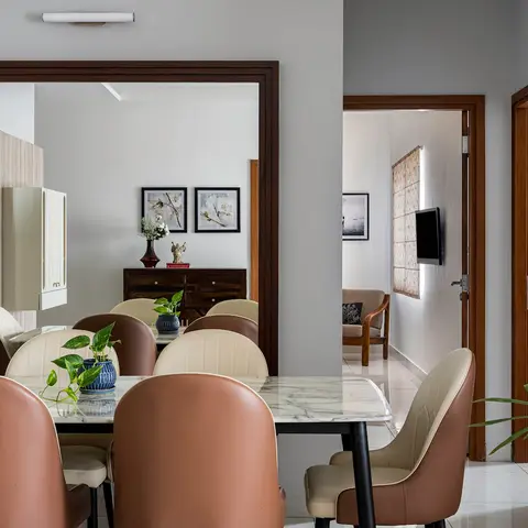 A dining room with a marble-top table and tan leather chairs reflected in a large wall mirror, a dark wood console with framed art beyond
