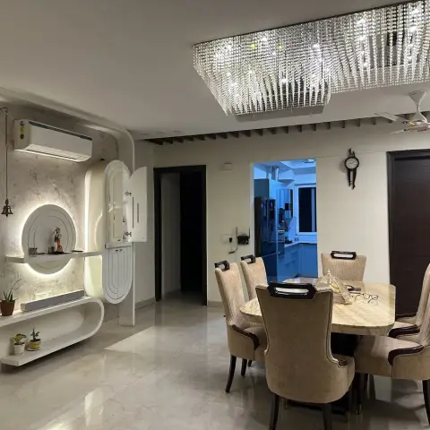 A dining room with a round marble table under a large crystal chandelier, beside a curved white backlit pooja niche wall.