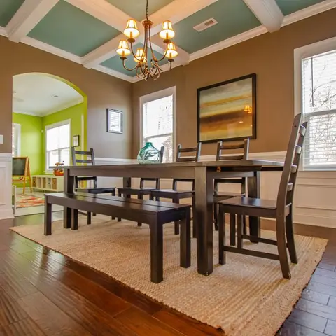 A dining room with a teal coffered ceiling and chocolate-brown walls with white wainscoting holds a dark wood table and bench under a candle chandelier.