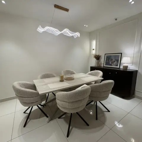 A dining room with a travertine-top table, six boucle swivel chairs, a sculptural wavy LED pendant light, and a dark wood console styled with framed art.