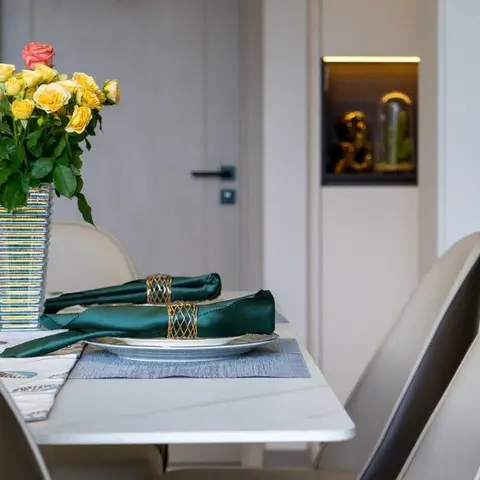 A dining table set with green napkins in gold rings and a bouquet of yellow roses, with beige leather chairs beside a backlit wall display niche.