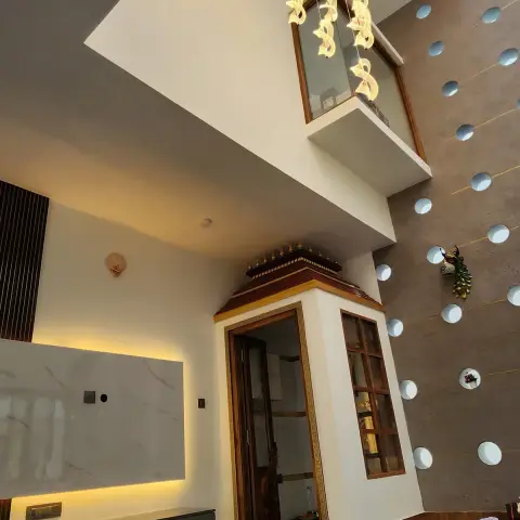 A double-height hall where a backlit white marble TV panel over a grey floating console faces a teak mandir with a carved crowned top and gold-beaded glass