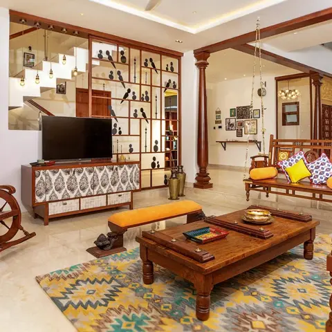 A double-height Indian living room with a carved wooden swing, rocking chair, and a low carved coffee table with brass diyas on a kilim rug