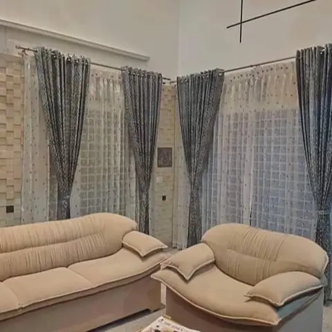 A double-height living room with 3D textured stone-tile wall panelling and floor-to-ceiling patterned sheer curtains behind a beige tufted rolled-arm sofa set.
