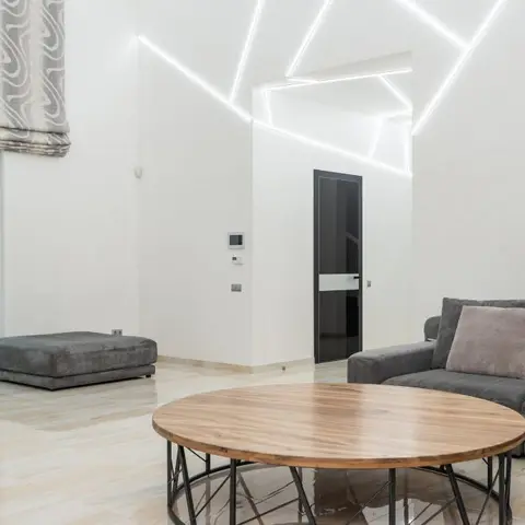 A double-height living room with angular white LED strips embedded in the ceiling above a round wood coffee table and grey sectional sofa.