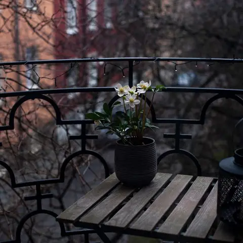 A dusk-lit balcony with an ornate black wrought-iron railing, a small wooden slat table holding a flowering potted plant and a lantern.