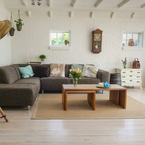A farmhouse-style living room with a white-painted exposed brick wall, exposed beam ceiling, a grey sectional sofa with bird-print cushions