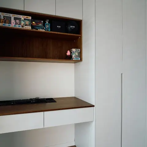 A floating walnut-top study desk beneath an open display shelf of collectibles, next to a plain white wardrobe wall.