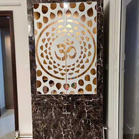 A floor-to-ceiling pooja cabinet with a laser-cut Om jaali door in dark marble-finish laminate, backlit to reveal deity photos inside.