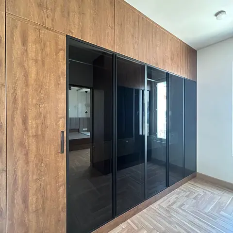 A floor-to-ceiling walnut wardrobe with black glass shutters reflecting a herringbone-floored bedroom with an upholstered headboard beyond.