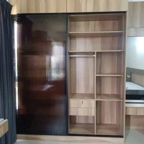 A floor-to-ceiling wardrobe with a fluted glass sliding shutter and open wood shelving, with a bed partly visible beyond it.