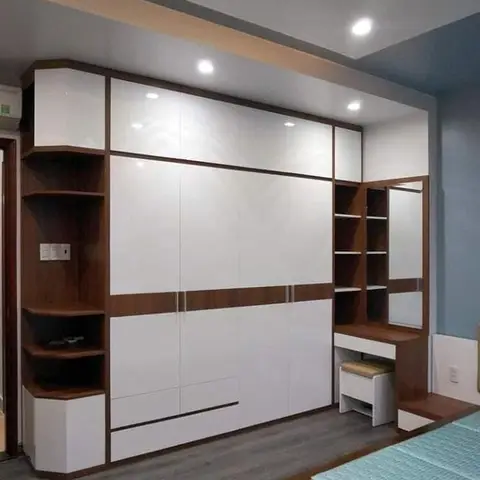A floor-to-ceiling white and wood-trim wardrobe wall with a built-in corner display shelf, an integrated dressing mirror and stool, and cove-lit ceiling above.