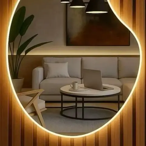 A fluted wood-panelled wall with an organic-shaped, LED-backlit mirror reflecting a grey sectional sofa, round coffee table