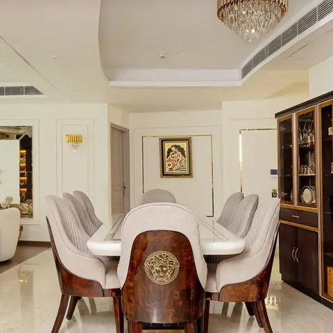A formal dining room with a marble-top table, brown wood chairs bearing a gold medallion emblem on the backs, a tiered crystal chandelier