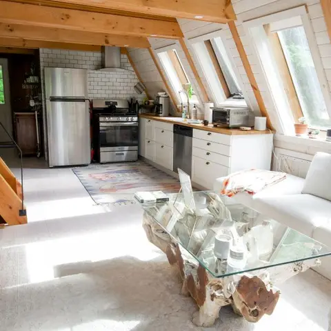 A-frame cabin living room with an off-white sectional sofa and a driftwood-base glass coffee table, exposed timber rafters, kitchen visible behind