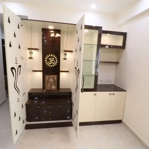 A freestanding pooja unit with laser-cut jaali doors in a tree-silhouette pattern, opening to reveal a backlit OM niche, brass diya lamps