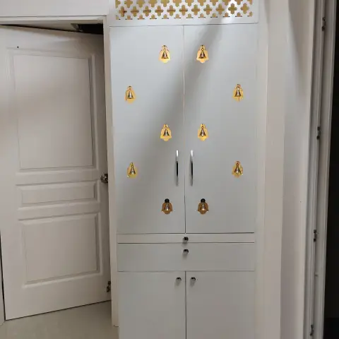 A freestanding white pooja cabinet with a gold cross-pattern jaali vent panel on top and rows of decorative brass bells adorning the closed doors