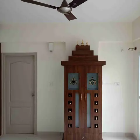 A freestanding wooden pooja unit with a tiered temple-shaped pediment, brass finials, hanging bells, and frosted glass door panels with traditional motifs.