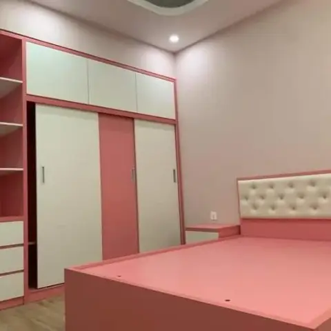 A girl's bedroom with a pink and white sliding-door wardrobe, an open corner display shelf, and a pink platform bed with a cream tufted headboard.