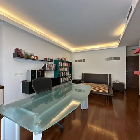 A glass-top study desk in the foreground with a cove-lit ceiling, wall-mounted bookshelves, and a low platform bed frame visible beyond.