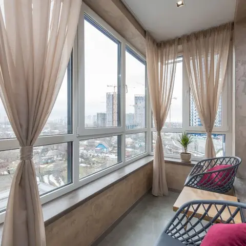 A glazed balcony with floor-to-ceiling windows, sheer beige curtains, a small wood table and woven bistro chairs with maroon cushions overlooking a city skyline
