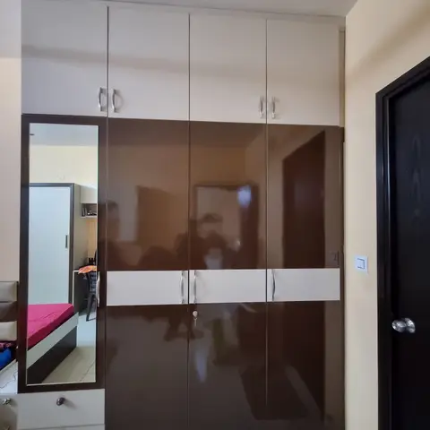 A glossy brown-and-cream wardrobe wall with a full-length mirror door reflecting an adjoining bedroom with a pink-sheeted bed.
