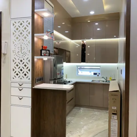 A glossy taupe modular kitchen with a marble breakfast counter, seen through a carved Ganesh-motif jaali partition door and backlit display shelves