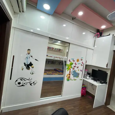 A glossy white sliding-door wardrobe printed with a footballer decal fronts a bunk bed reflected in its mirror, under a pink-and-blue playful pop ceiling.