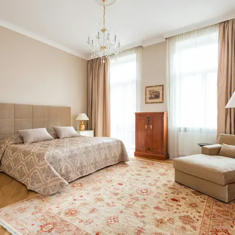 A grand master bedroom with a crystal chandelier, floor-length silk drapes, an upholstered headboard, a chaise lounge, and a large antique-pattern rug