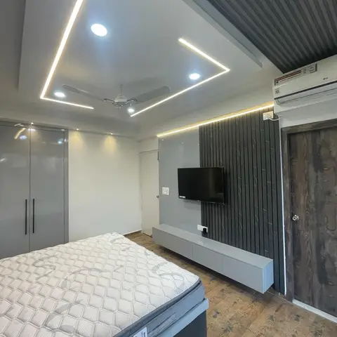 A grey bedroom with a glossy wardrobe wall and a wall-mounted TV on dark fluted panelling under a cove-lit rectangular false ceiling