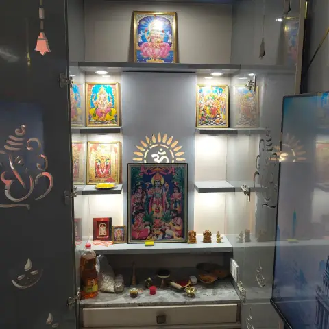A grey lacquered pooja cabinet with laser-cut Om and diya motif doors, backlit niches displaying framed deity portraits and brass idols.